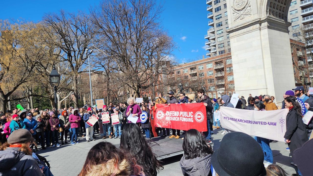 A speaker addresses the crowd at the Stand Up For Science rally while people hold large banners that read "Defend Public Research Funding"