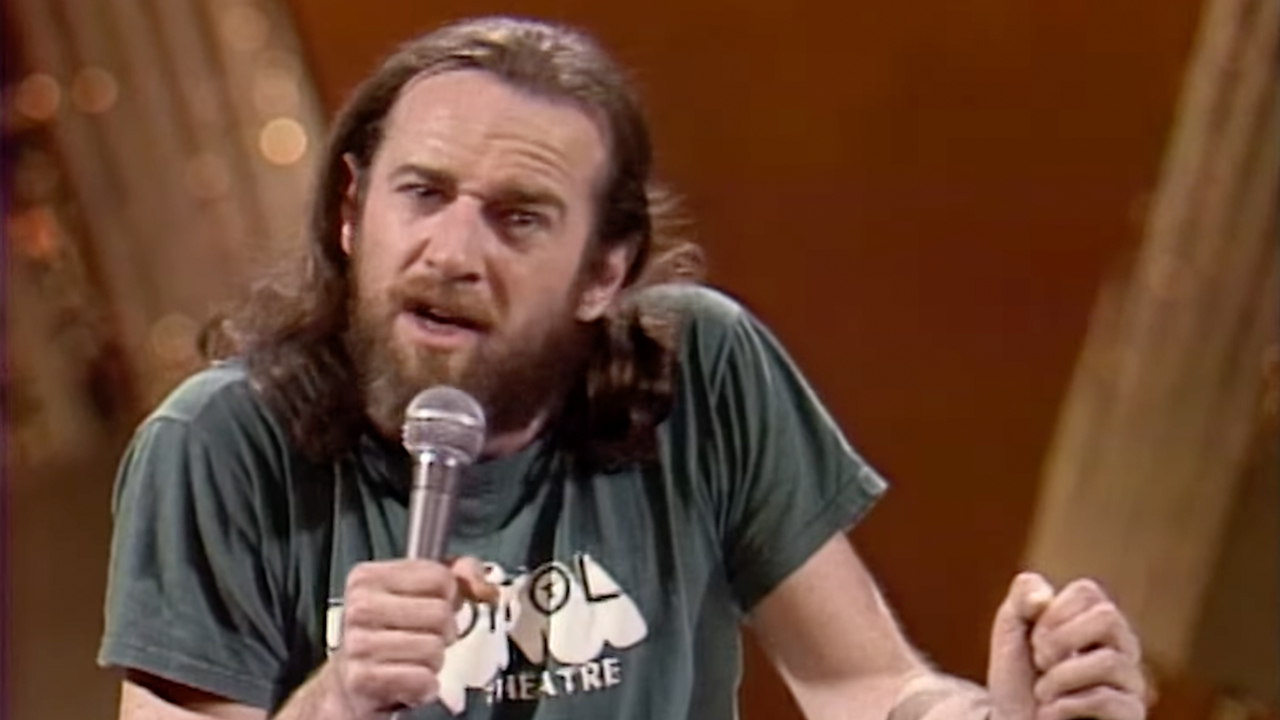 Archival footage of George Carlin with long hair and a theater shirt performing stand up, used in HBO's doc George Carlin's American Dream.