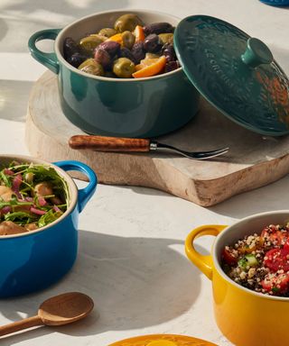 Potato salad in a blue cocotte; a quinoa, cucumber, and tomato salad in a yellow cocotte; olives and oranges in a green cocotte; all on a white countertop.