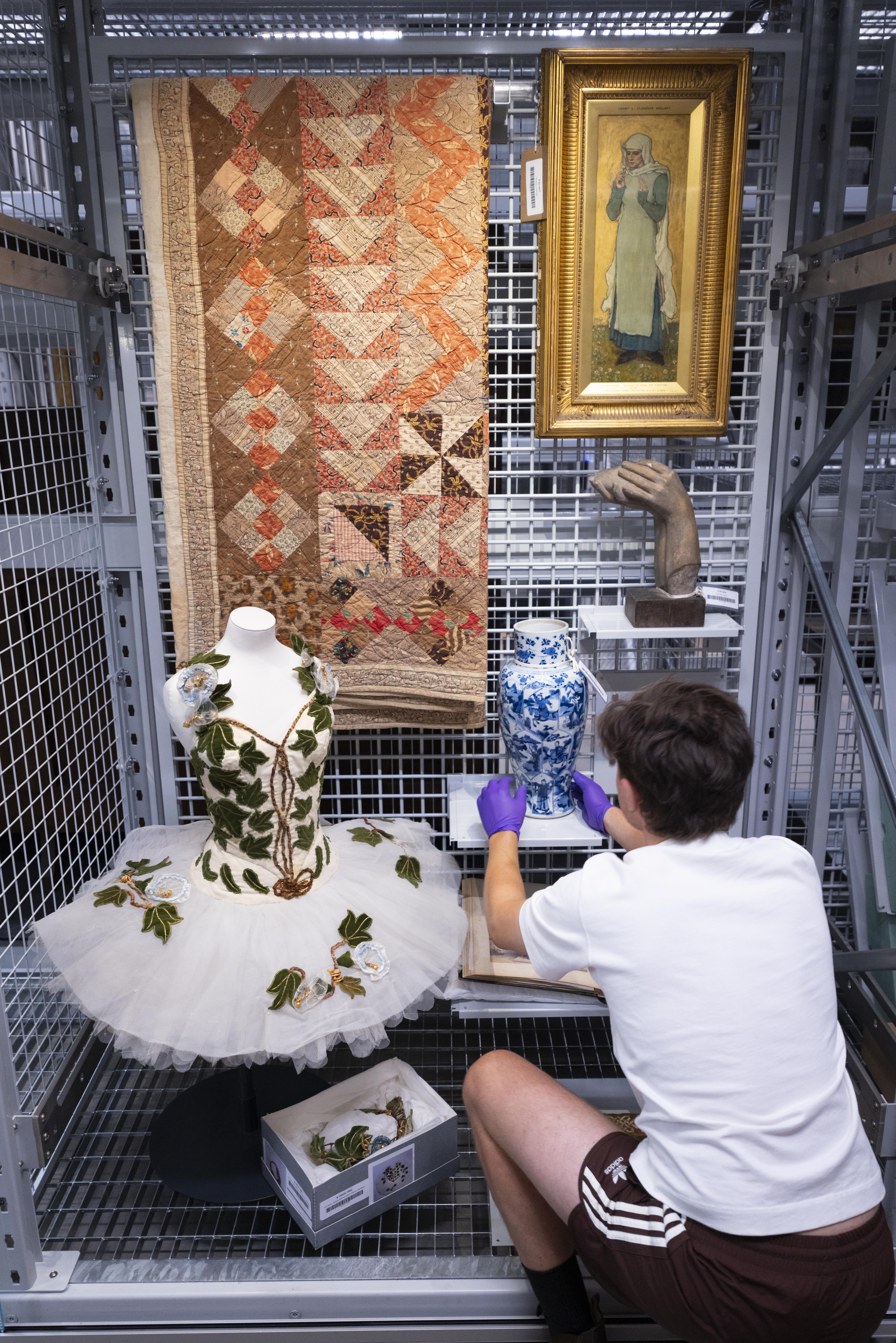 A man in a white t-shirt arranges museum artefacts, including a wide tutu strewn with ivy leaves, in a metal grill display case