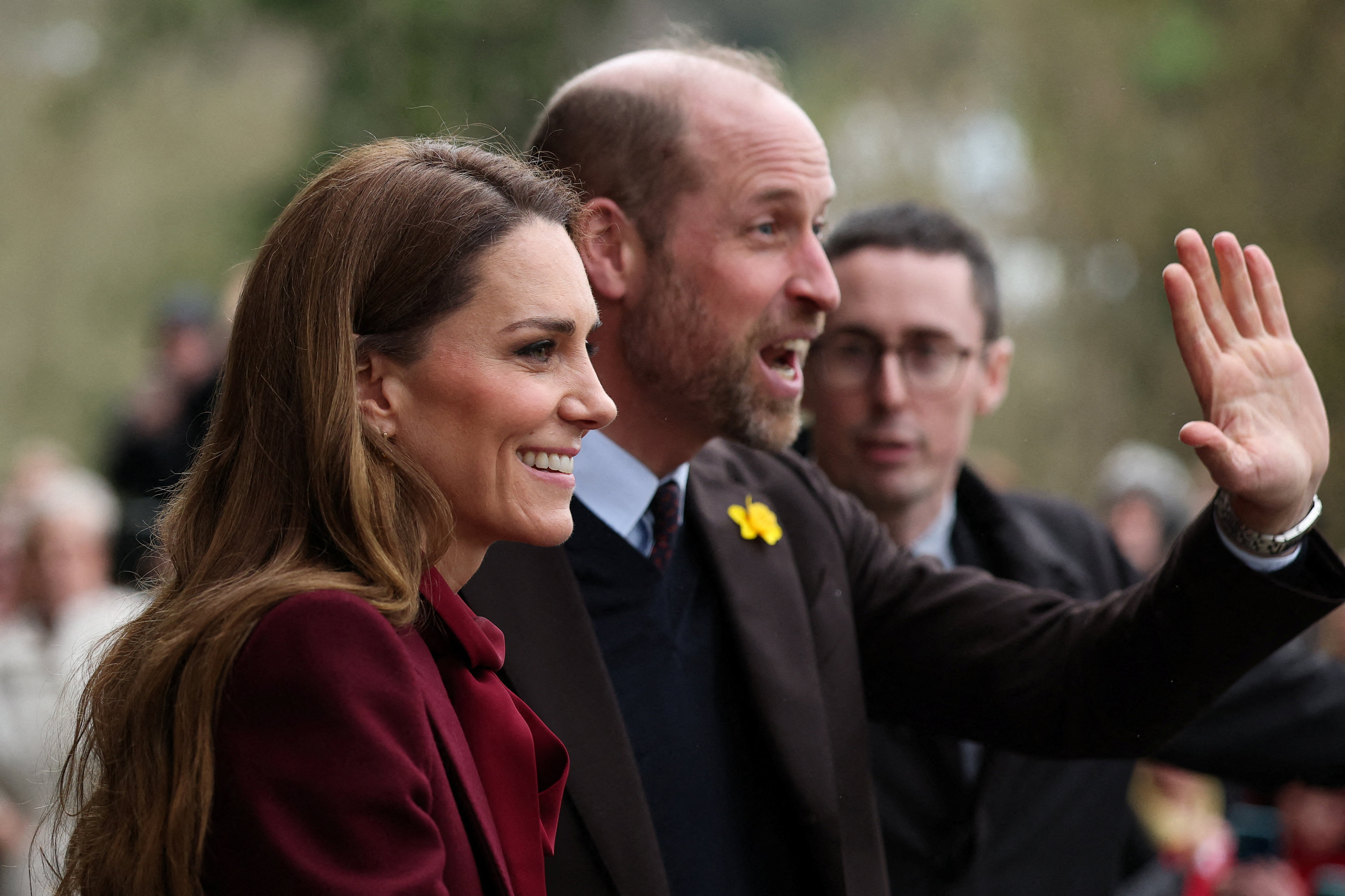 Prince William standing next to Princess Kate, wearing a red coat, and waving