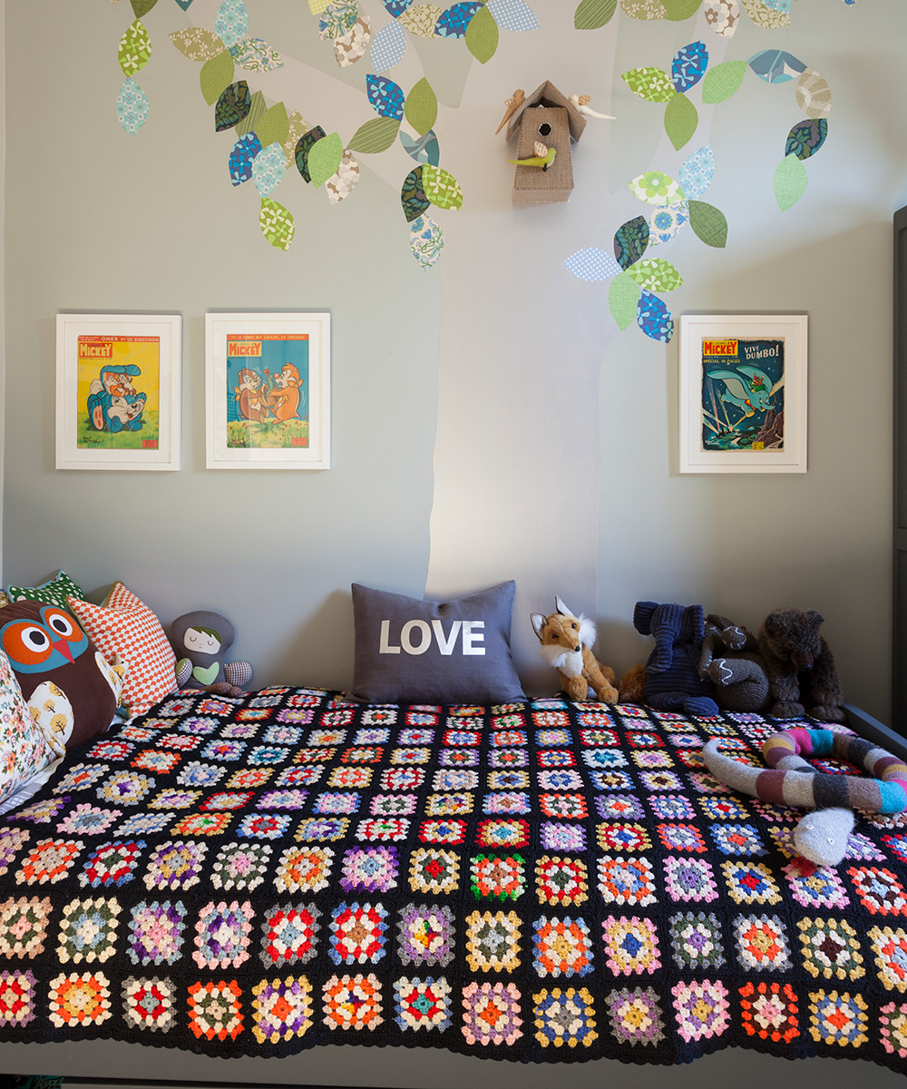 Small bedroom ideas for kids illustrated by wall art on a pale green-gray wall behind a bed with a crochet quilt and an assortment of cushions and stuffed toys.