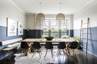 dining room with slate gray walls painted halfway up, stone painted upper walls and ceiling, dark wood floor, rustic dining table, Eames black chairs, artwork, animal rug, rattan shade, bench