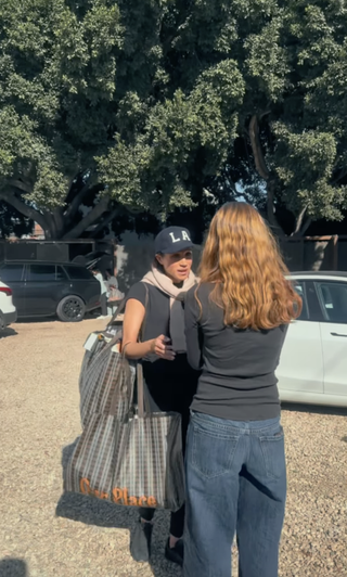 Meghan Markle wearing an LA baseball cap and a black tee shirt carrying shopping bags and talking to a girl wearing jeans and a black tee with her backt o the camera in front of two cars.