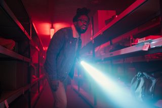 Nick (Adjani Salmon) stands in a storage unit lit by red light, holding a bright torch and looking toward the camera