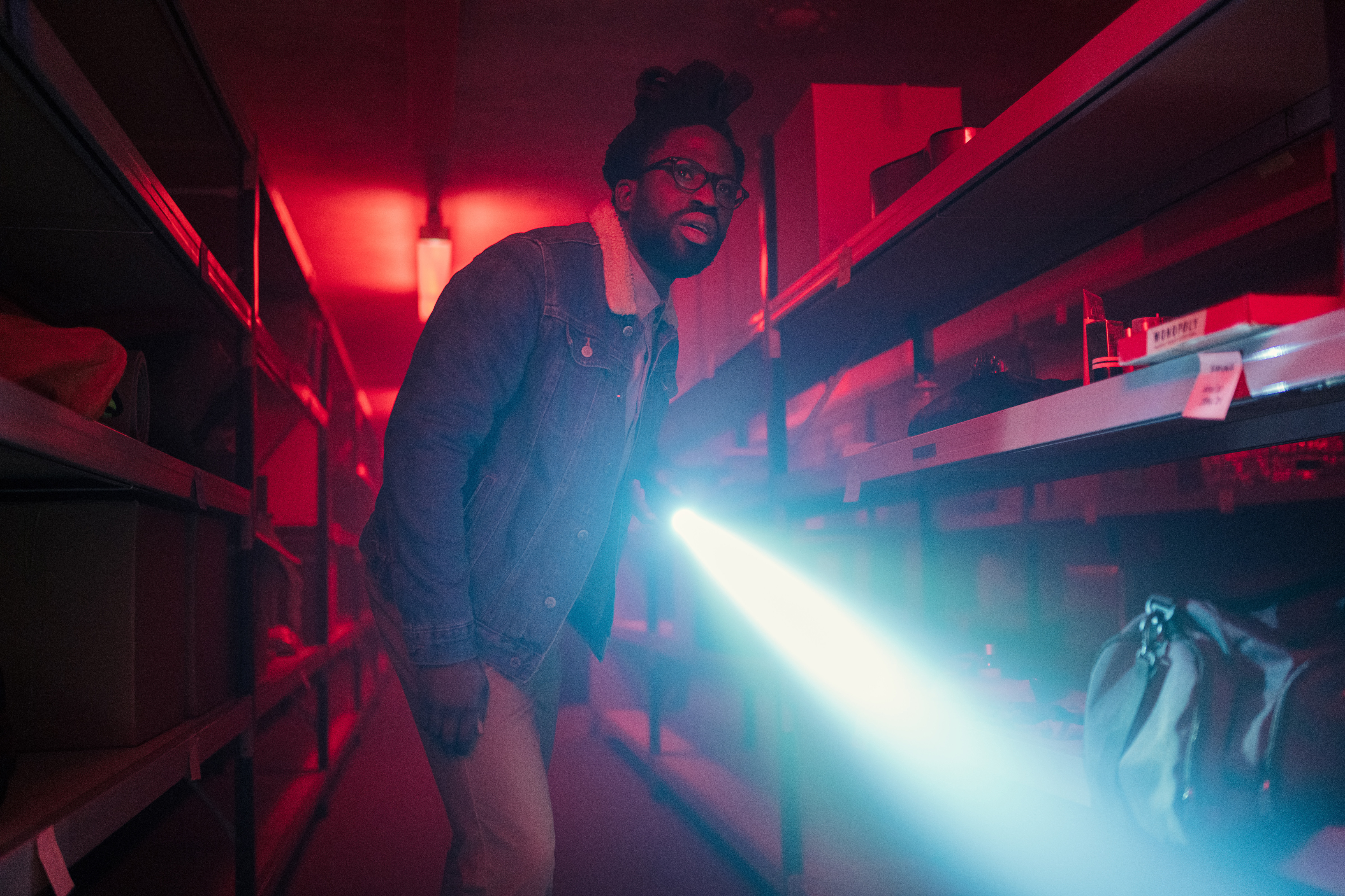 Nick (Adjani Salmon) stands in a storage unit lit by red light, holding a bright torch and looking toward the camera