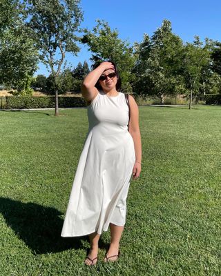 fashion influencer Marina Torres poses in a field of grass wearing a simple sleeveless white midi dress, sunglasses, and flip-flop sandals