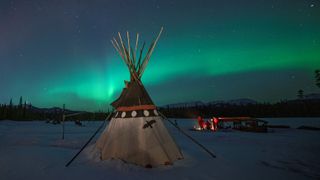 A teepee in the center of the image with a starry sky above and the aurora borealis stretching across the sky in a glowing green ribbon of light. 