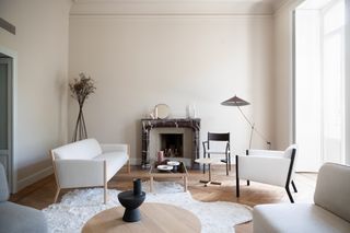 A room with simplistic furniture. A white sofa with wood trim. A white chair with black trim. A grey chair with a white base. A fireplace with candles and a mirror on top. A small wood table with tableware. Ariake installation Milan