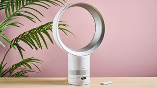 The Dyson Cool CF1 is facing slightly to the right on a table top in front of a leafy green plant and a pink wall. The remote control is on the table to the right of the fan.