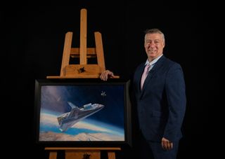 a man in a suit smiles beside a painting on an easel. the painting depicts a triangular space plane and a satellite above Earth