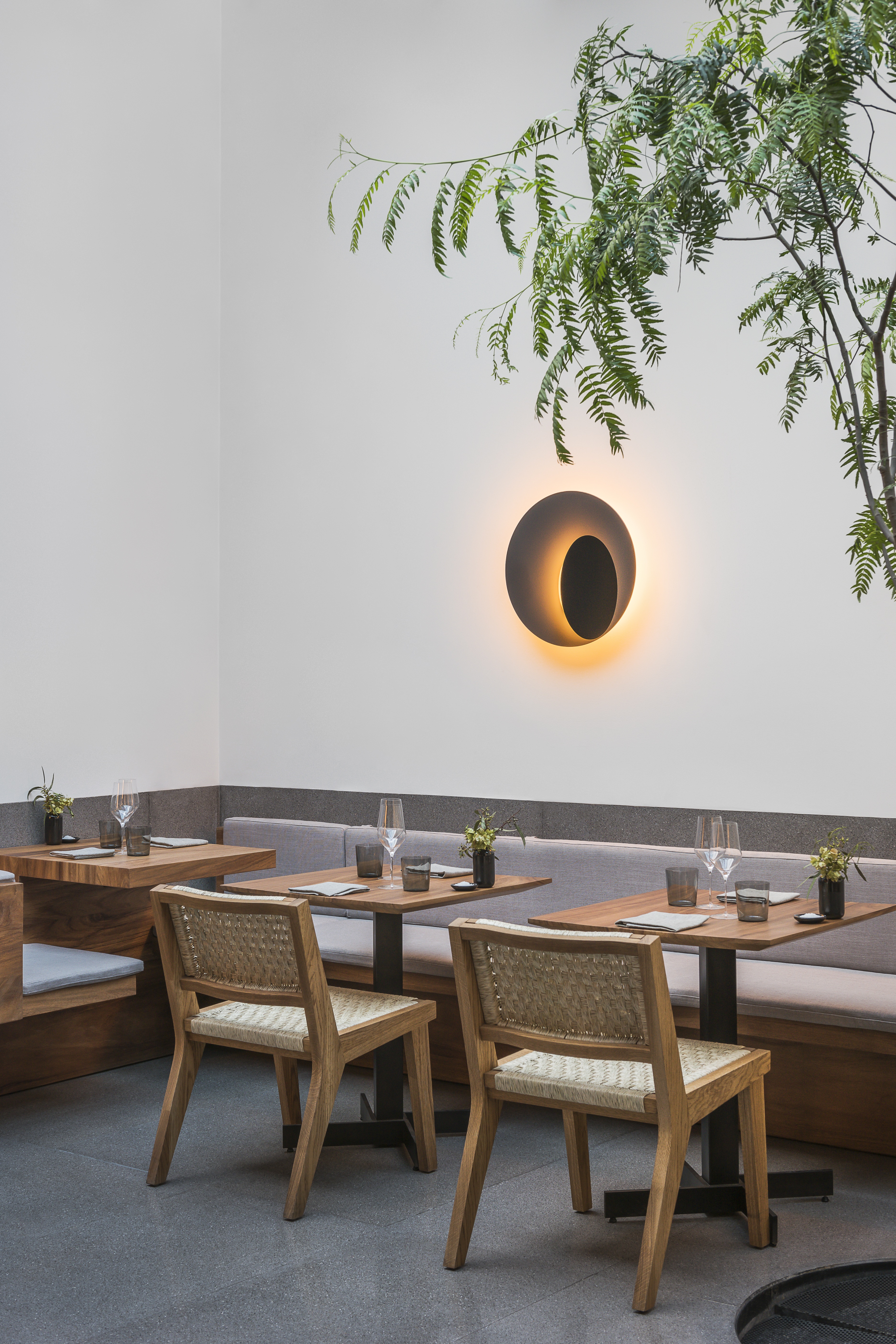 A modern rustic-meets-brutalism restaurant features rattan and wood chairs, upholstered banquette in a faint shade of grey, spherical sconces mimicking the sun, grey floors, and plants.