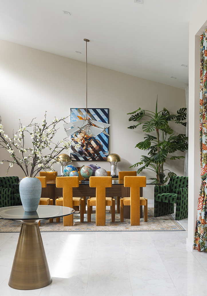 dining room with sloped ceiling, white walls and ceiling, timber dining table with mustard yellow dining chairs, artwork on wall, tall monstera plant in the corner, console table, hanging pendant over dining table, rug under dining table, and console table in the foreground with a vase and flowers