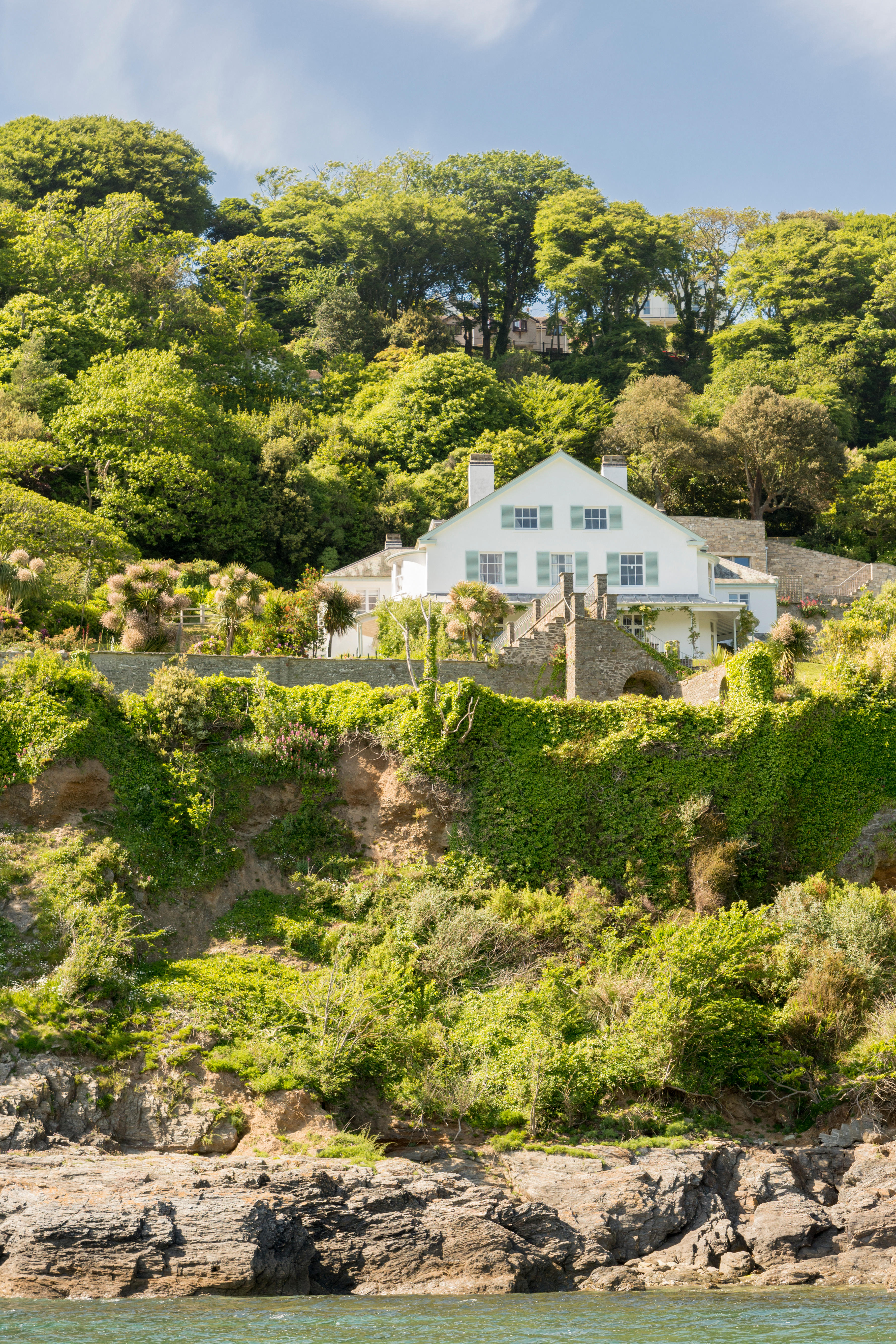 A white house with blue shutters on a steep, tree-covered cliff, overlooking the water