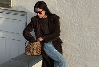 Annabel wears a brown suede trench coat, brown shirt, jeans, and a leopard bag.