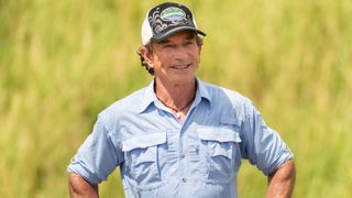 Jeff talks to the contestants ahead of the third challenge on Survivor. He's wearing a blue button-up shirt, smiling, and has a background of grass behind him. 