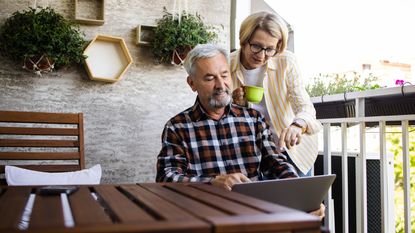 Portrait of a happy senior couple enjoying coffee on their balcony, using laptop and making some plans together