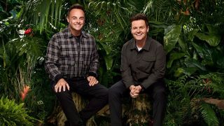 Against lush green foliage, hosts Ant McPartlin and Declan Donnelly sit on tree stumps sporting Cheshire cat grins, anticipating the arrival of the latest celebrity campmates for "I'm a Celeb" 2025.