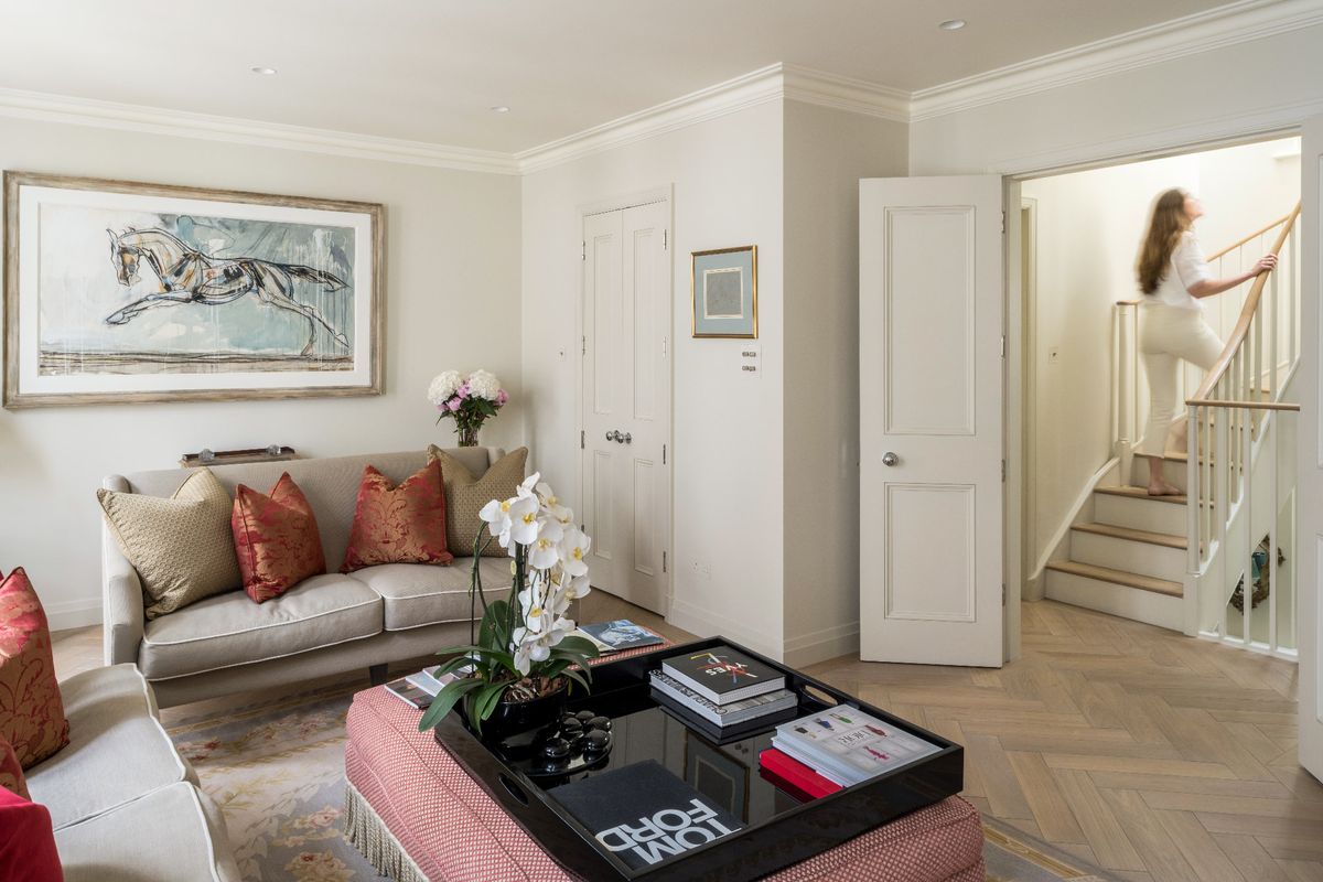 A living room with a plush beige sofa topped with red and beige cushions, a woman descends a staircase to the back of the room