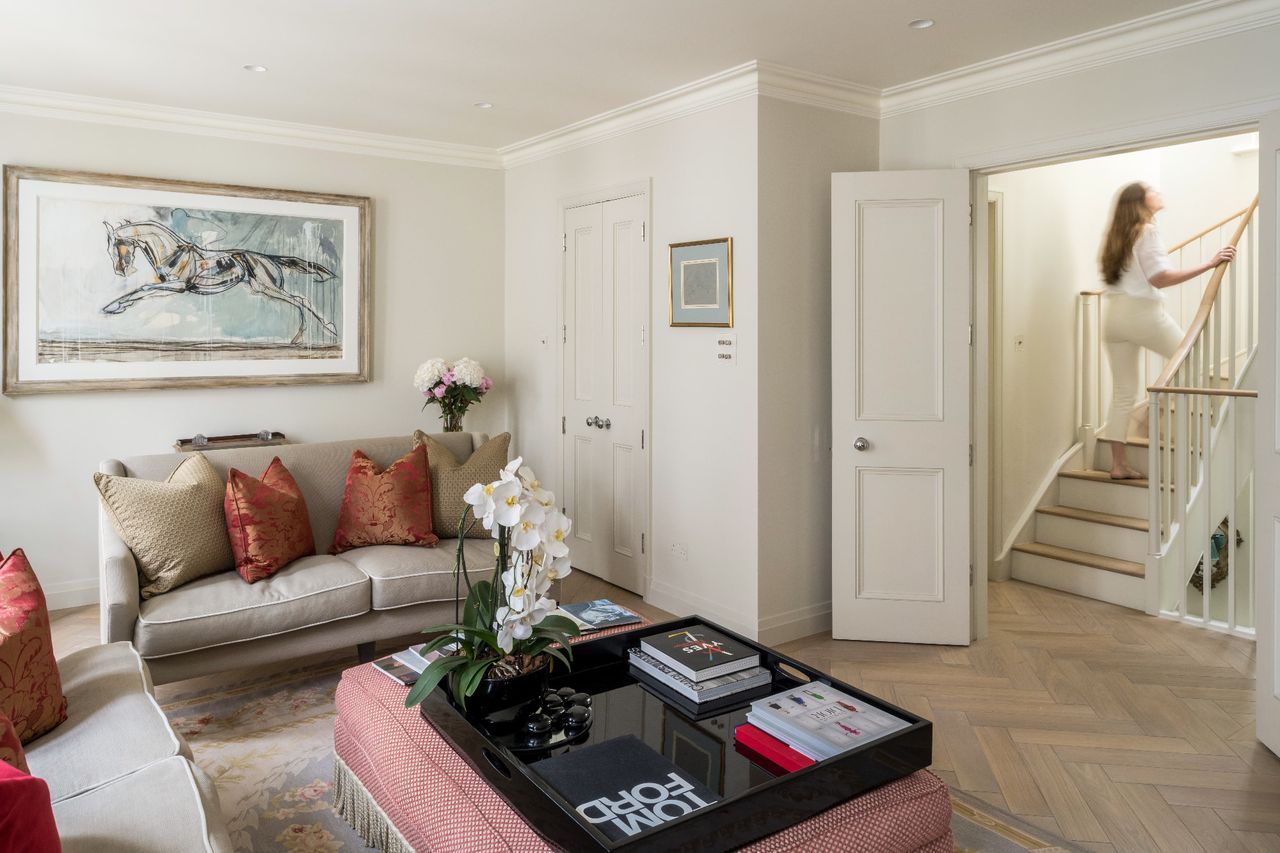A living room with a plush beige sofa topped with red and beige cushions, a woman descends a staircase to the back of the room