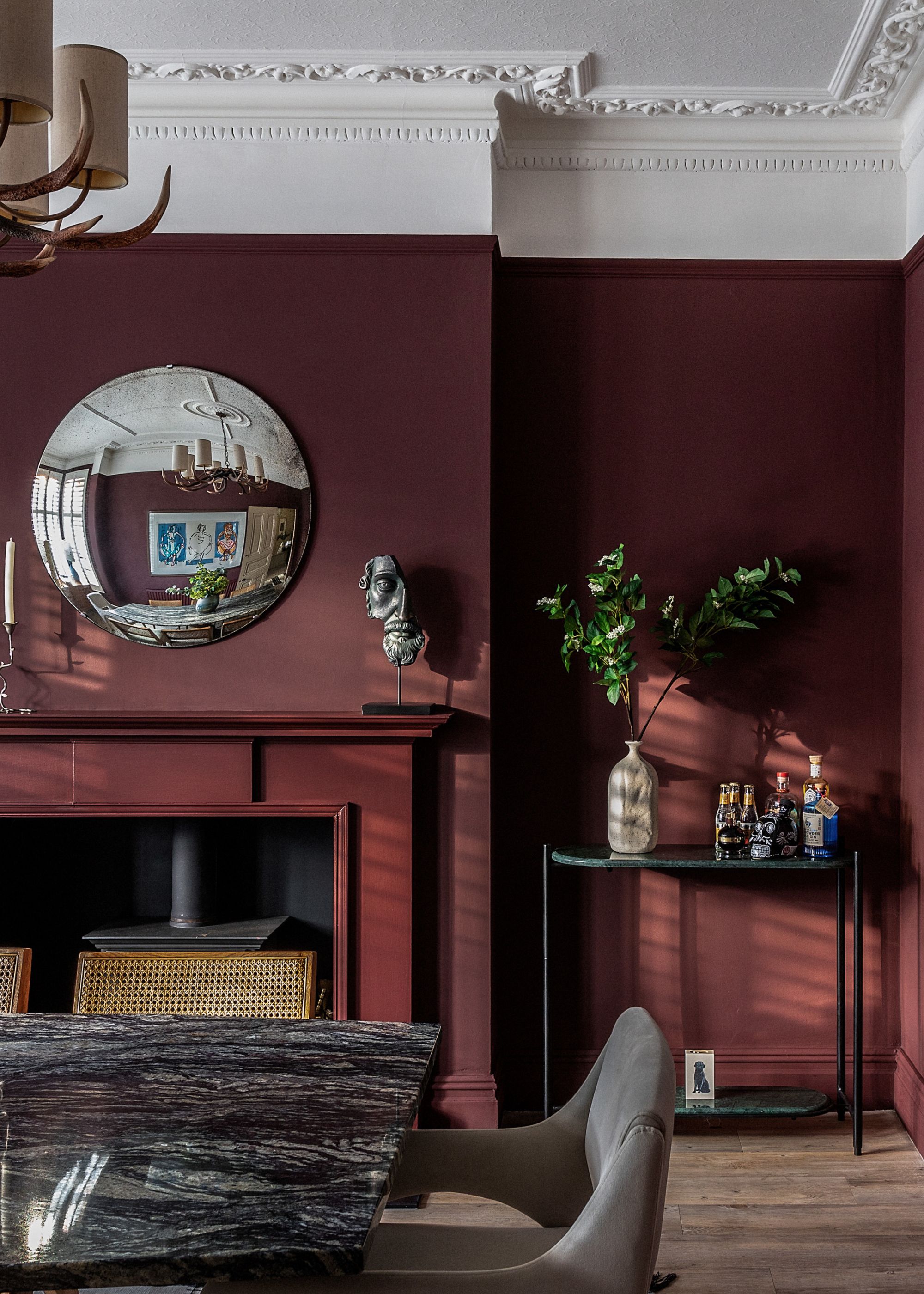 A burgundy dining room with white ceilings and moldings, a stone dining table, a convex mirror above a fireplace next to a small bar table with foliage in a vase
