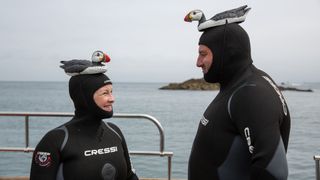 Wynne and Joanna standing on a boat in wetsuits both with plastic puffins strapped to their heads