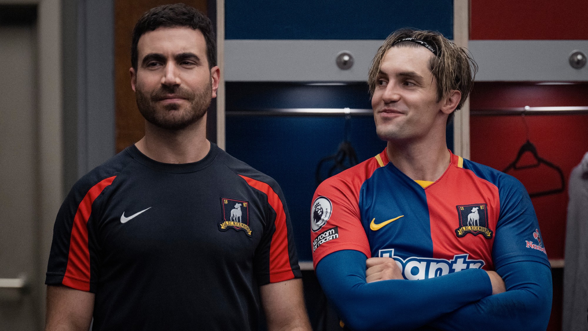 From left to right: Brett Goldstein as Roy Kent almost smiling and Phil Dunster as Jamie Tartt smiling and looking at Roy in Ted Lasso.
