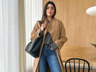 British fashion influencer Anna Howard poses in her chic wood-paneled London kitchen wearing a camel coat, gray sweater, black tote bag, black belt, and jeans.