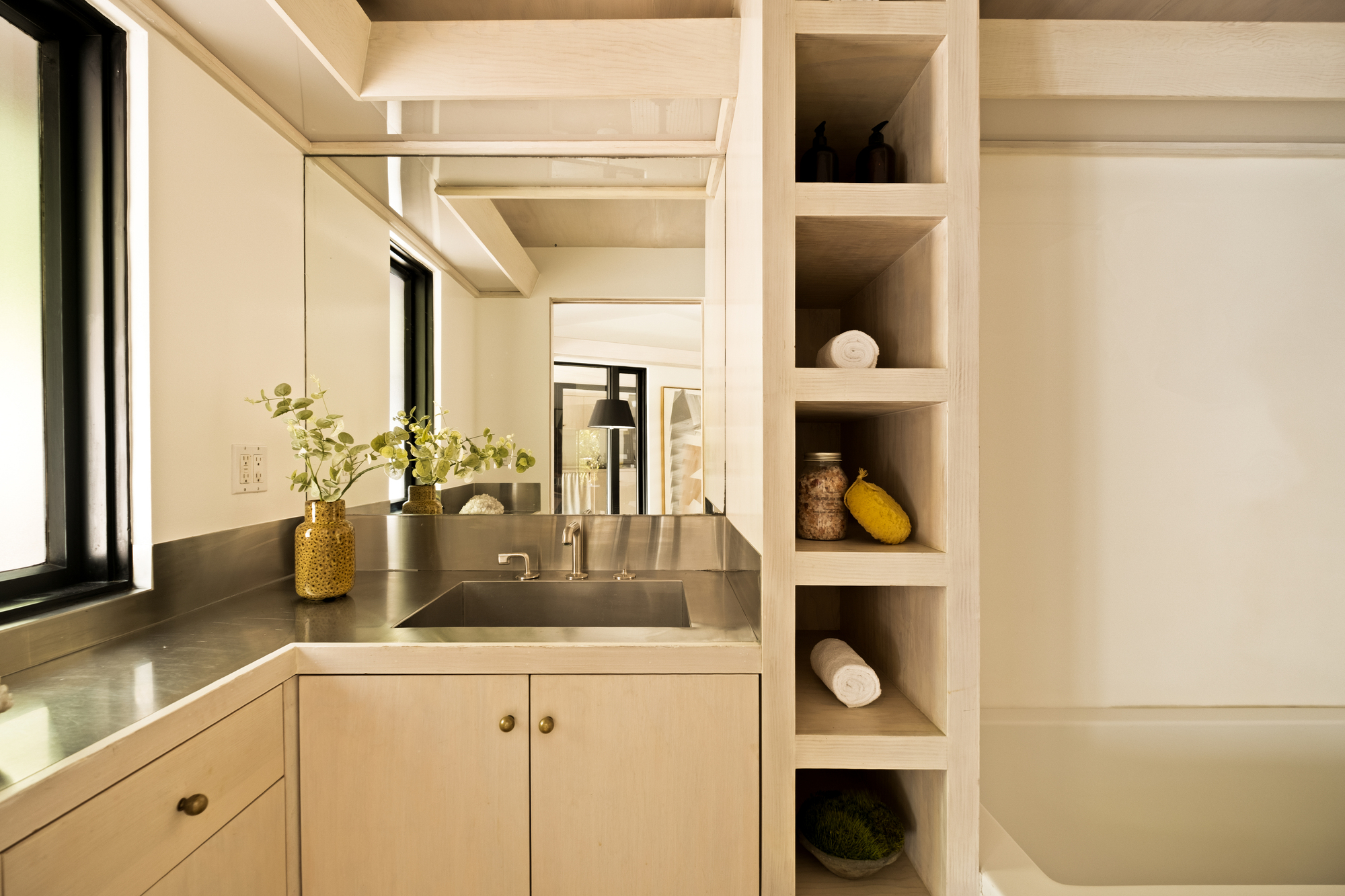 Image of a bathroom vanity area that has stainless steel countertops and off-white cabinetry. There is floor-to-ceiling shelving and a tub in view.