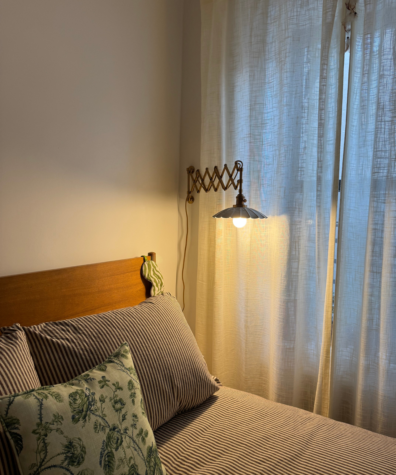 a bedroom with ivory curtains, a wooden bed frame, striped bedding, and a plug in wall sconce