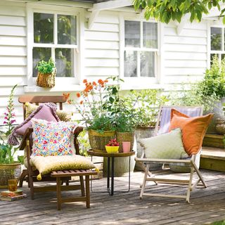two deckchairs with colourful cushions on decking by the white wooden clad house with pots of plants