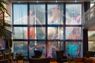 The aerial shot of a group of friends bathing at sea, depicted from above, has been turned into a textile print made of three separate pieces of fabric, now hanging in a dimly lit hotel lobby.