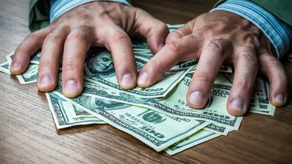 A man's hands appear to grab a pile of hundred-dollar bills on a table.
