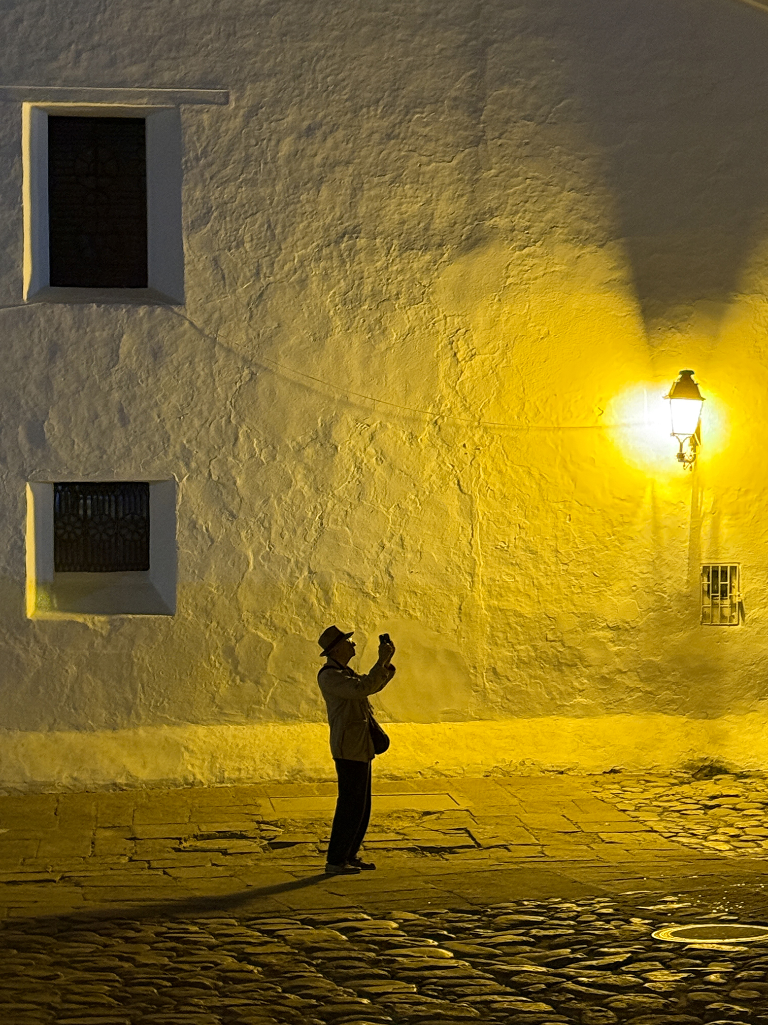 A person in a hat takes a photo in front of a softly lit white wall, illuminated by a vintage street lamp at night