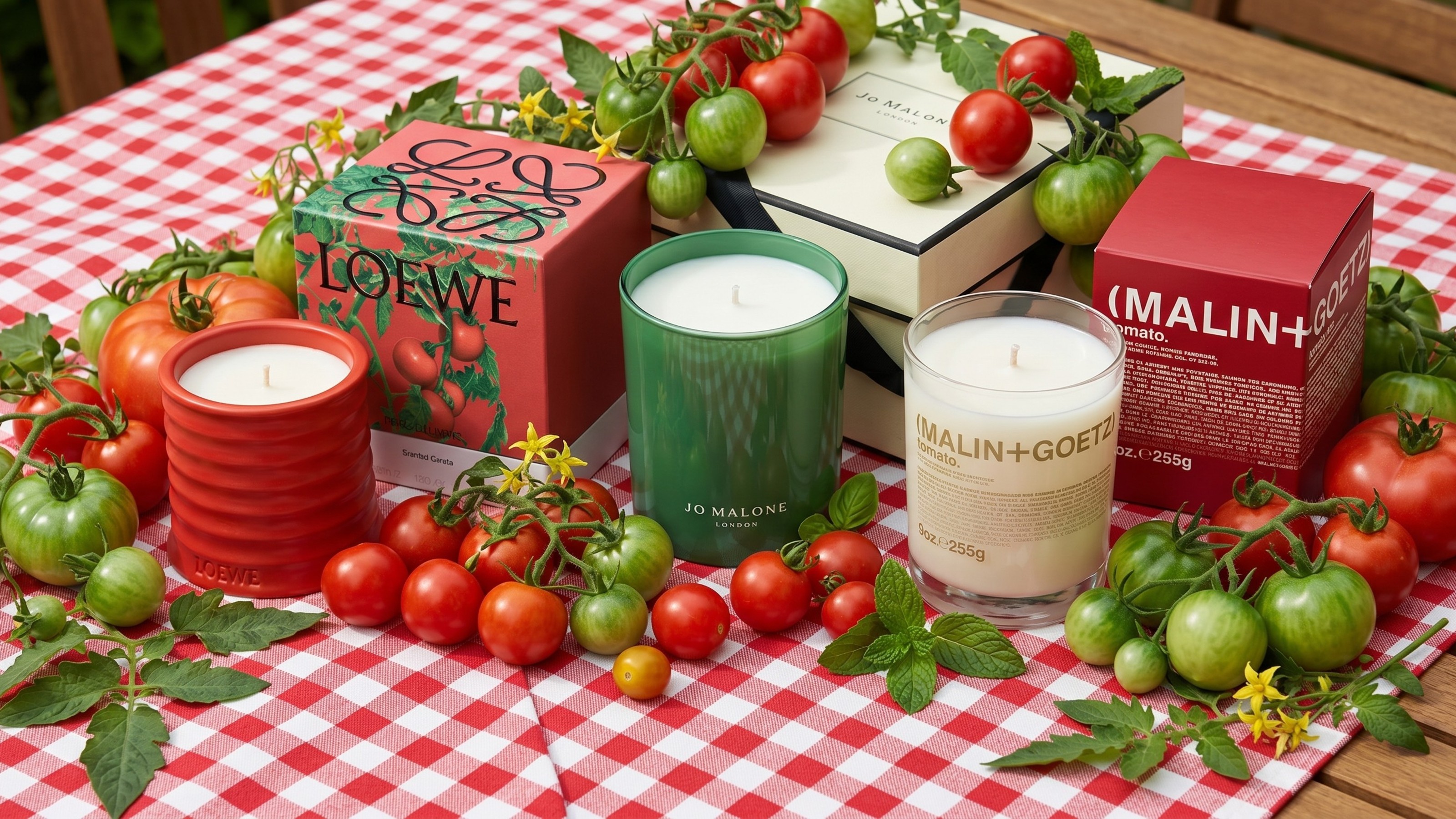 A table with a red gingham tablecloth and tomato-scented candle from Loewe, Jo Malone and Malin + Goetz surrounded by red and green tomatoes