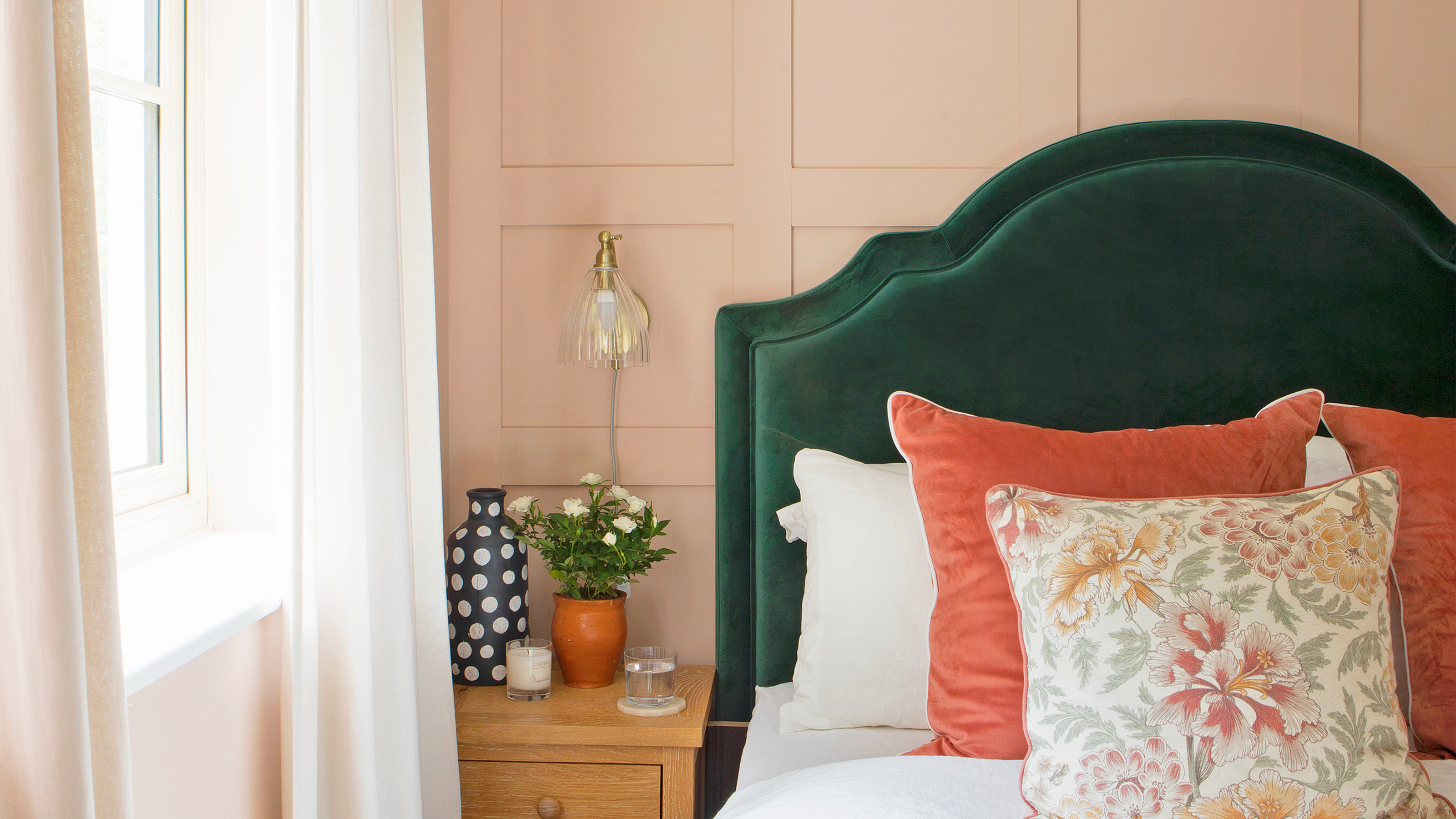 A bedroom with a panelled wall painted in peach paint, and a bed with a green upholstered headboard and orange cushions