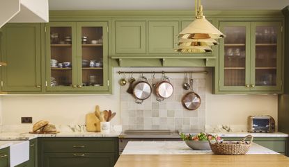 A double tones green kitchen with gold pendant lights hanging above a kitchen island. There is also a view of the stove where pots and pans hang above it
