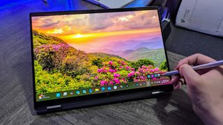A person using a stylus on a HP OmniBook X Flip 14 (2025) laptop displaying a vibrant sunset over a mountain landscape with blooming pink flowers, creating a serene atmosphere.