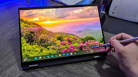 A person using a stylus on a HP OmniBook X Flip 14 (2025) laptop displaying a vibrant sunset over a mountain landscape with blooming pink flowers, creating a serene atmosphere.