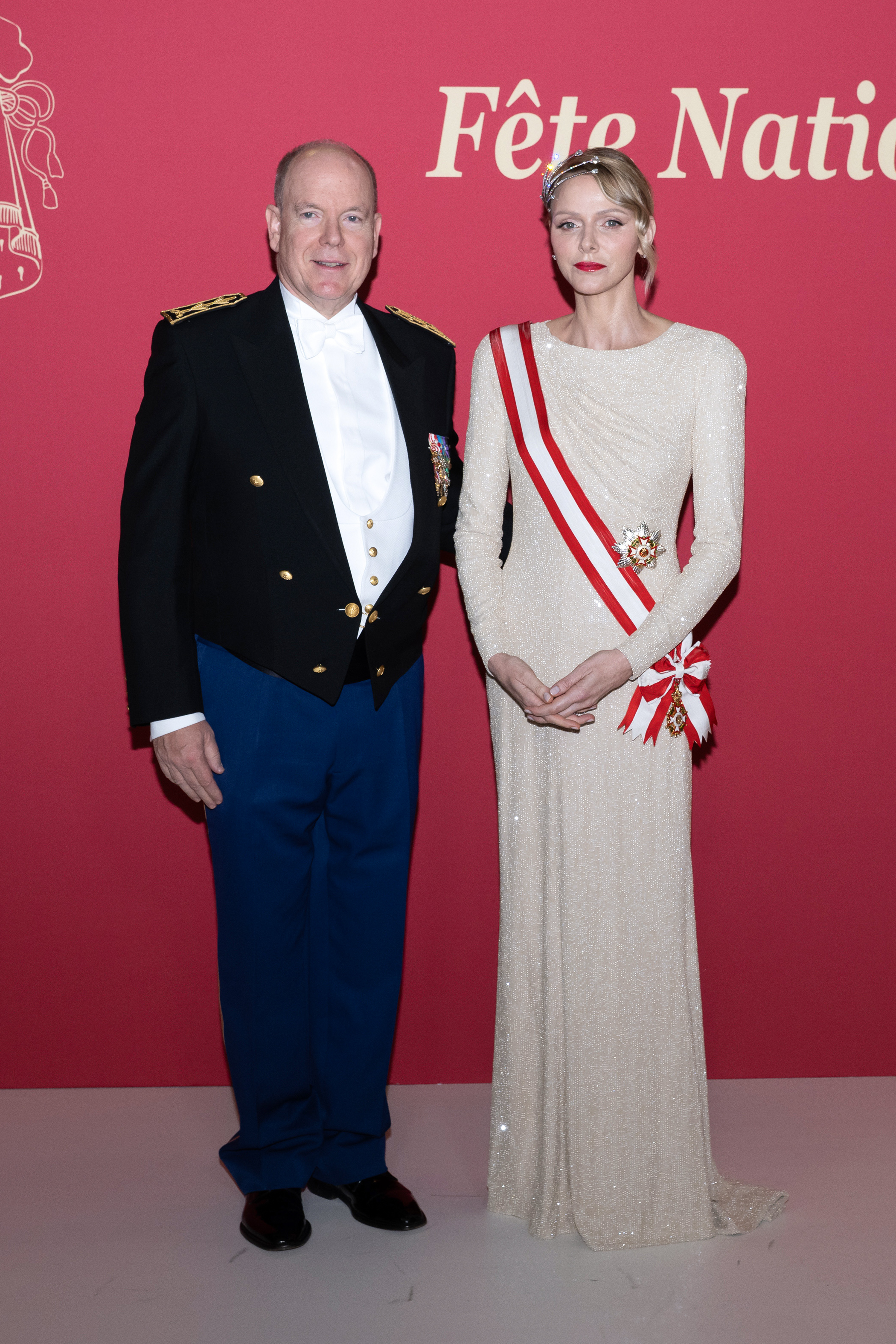 Princess Charlene wearing an ivory long sleeved dress and red and white sash standing with Prince Albert on a red carpet