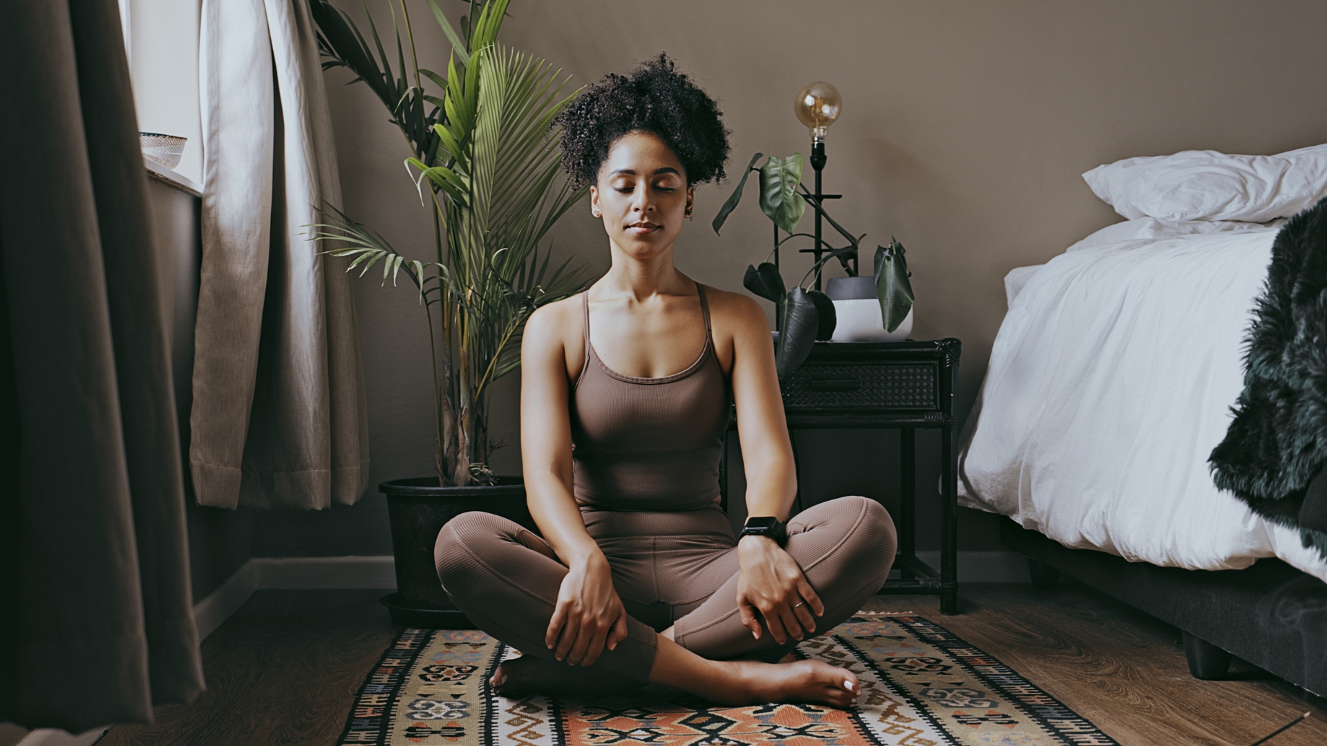 woman in brown leggings and vest sat on a rug in a bedroom next to a bed with white sheets with a bedside table and plant behind her and window the other side of her.