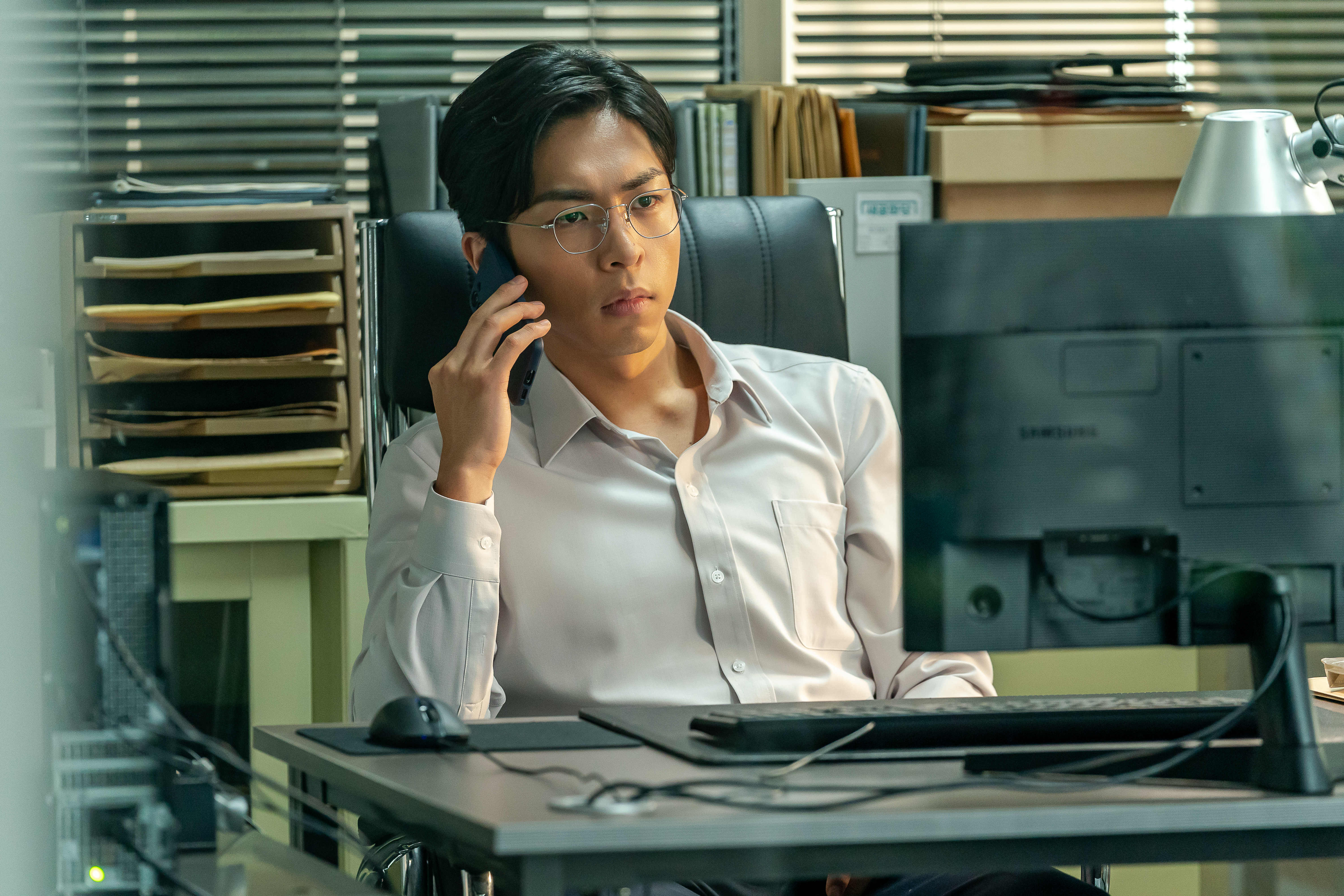 Joo Jong-hyuk sits at a desk in front of a desktop computer on the phone in &#039;Tempest.&#039;