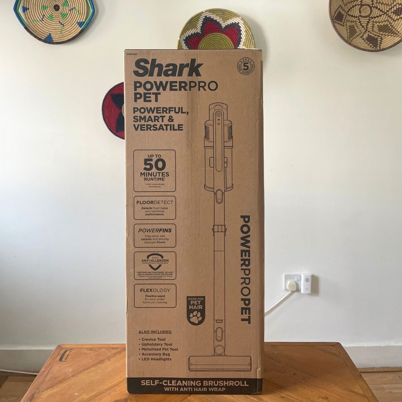 Shark PowerPro Pet Cordless Stick Vacuum box sitting on a wooden table in front of a white wall with colourful baskets on it