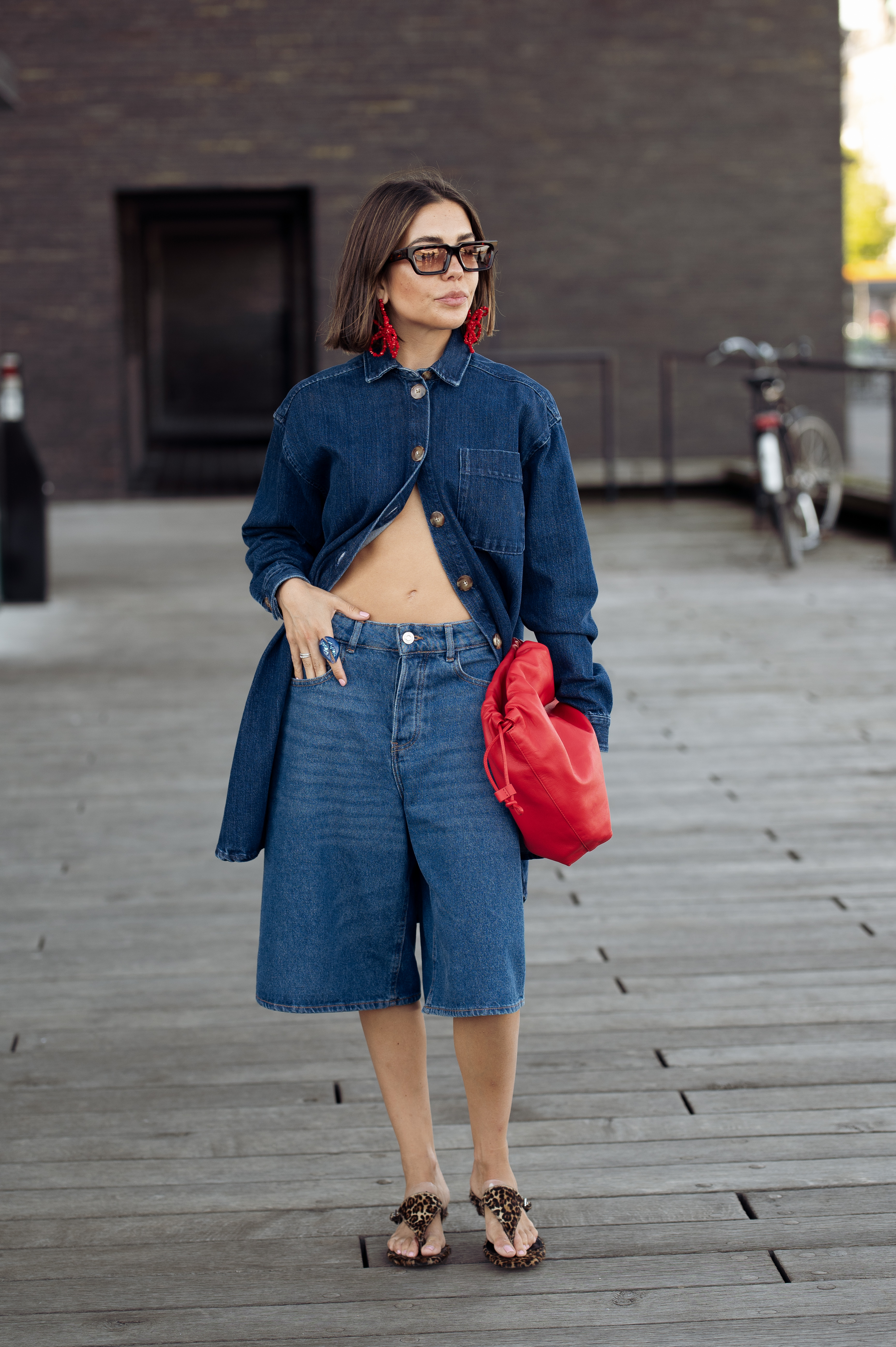 woman wears jeans, blue denim shirt, brown flip-flops, red bag and sunglasses on August 05, 2024 in Copenhagen, Denmark.