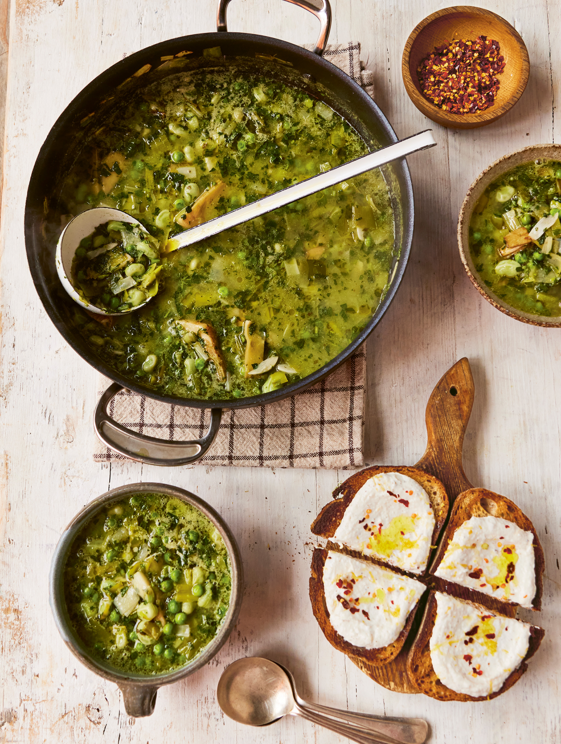 A pan full of green soup accompanied by a wooden board laden with toast and ricotta cheese
