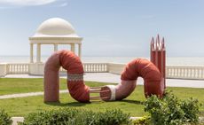 Holly Hendry Invertebrate art exhibit . A Waterfronts commission with the De La Warr Pavilion. A red worm like structure on a green lawn with a sea view.
