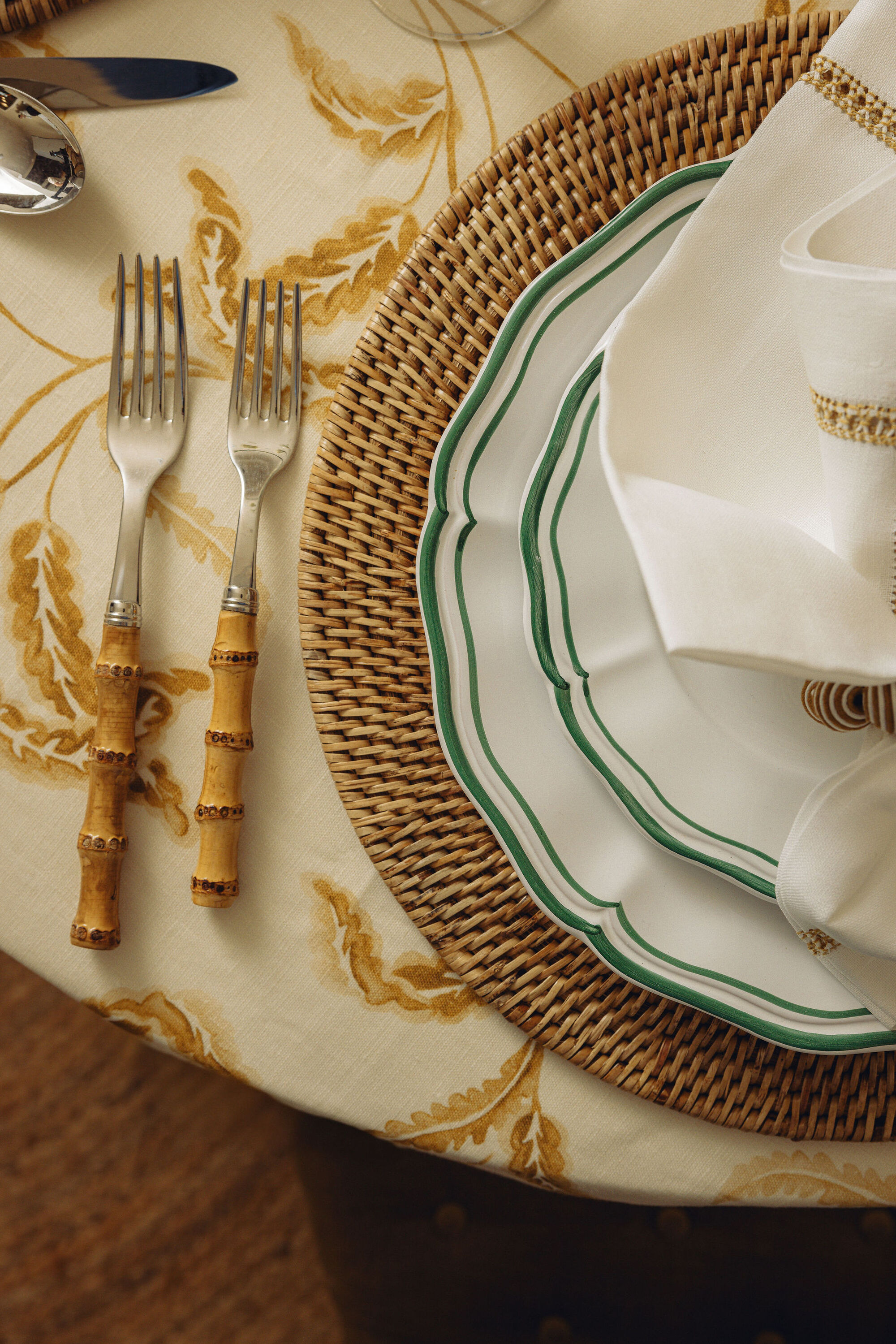 Close-up of a table set with a white and yellow floral tablecloth, bamboo cutlery, a wicker charger plate, and green and white dinner plates.