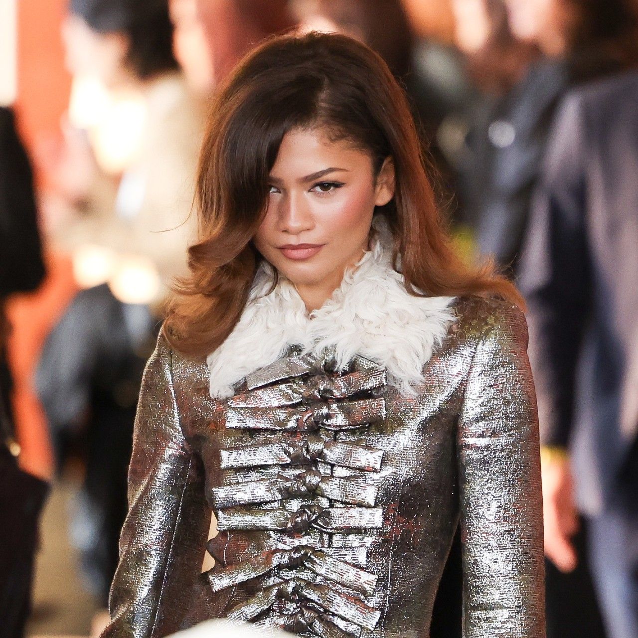 a woman with brown hair posing for a camera while wearing a metallic jacket with feather trim