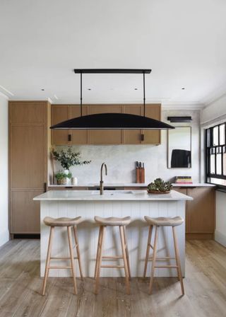 Oak/wood beadboard kitchen cabinet with a white paneled kitchen island and cream-colored bar stools. Above the kitchen island is a large pendant light.