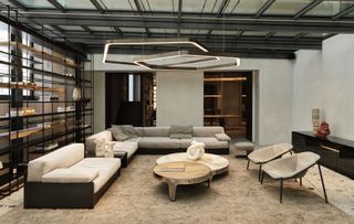 A large white and grey L shaped sofa with two low-back chairs surround two wood circular tables. Two hexagonal light shades hang from the ceiling. A large metal freestanding shelving unit runs along the left wall.